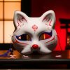 Masque japonais Kitsune blanc en forme de renard avec motifs rouges et bleus, cordon violet, posé sur une surface sombre devant un décor traditionnel japonais