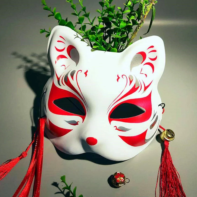 Masque japonais Kitsune en céramique blanche avec motifs rouges traditionnels, décoré de pompons rouges et suspendu devant une plante verte