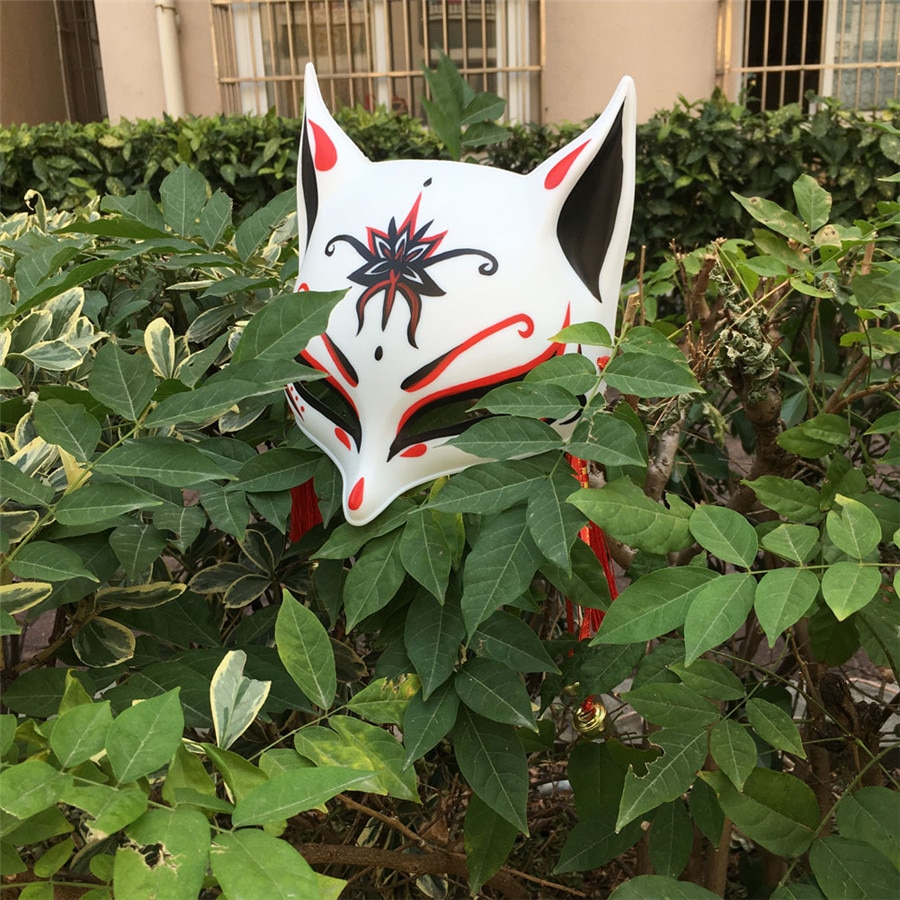 Masque japonais Kitsune Le Retord en plastique blanc avec motifs rouges et noirs, décoré d'un motif rouge et noir sur le front, posé dans des feuillages verts